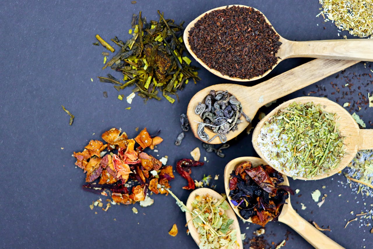 hero-img-01 Top view of various herbal tea blends displayed on wooden spoons, showcasing a mix of textures and colors.
