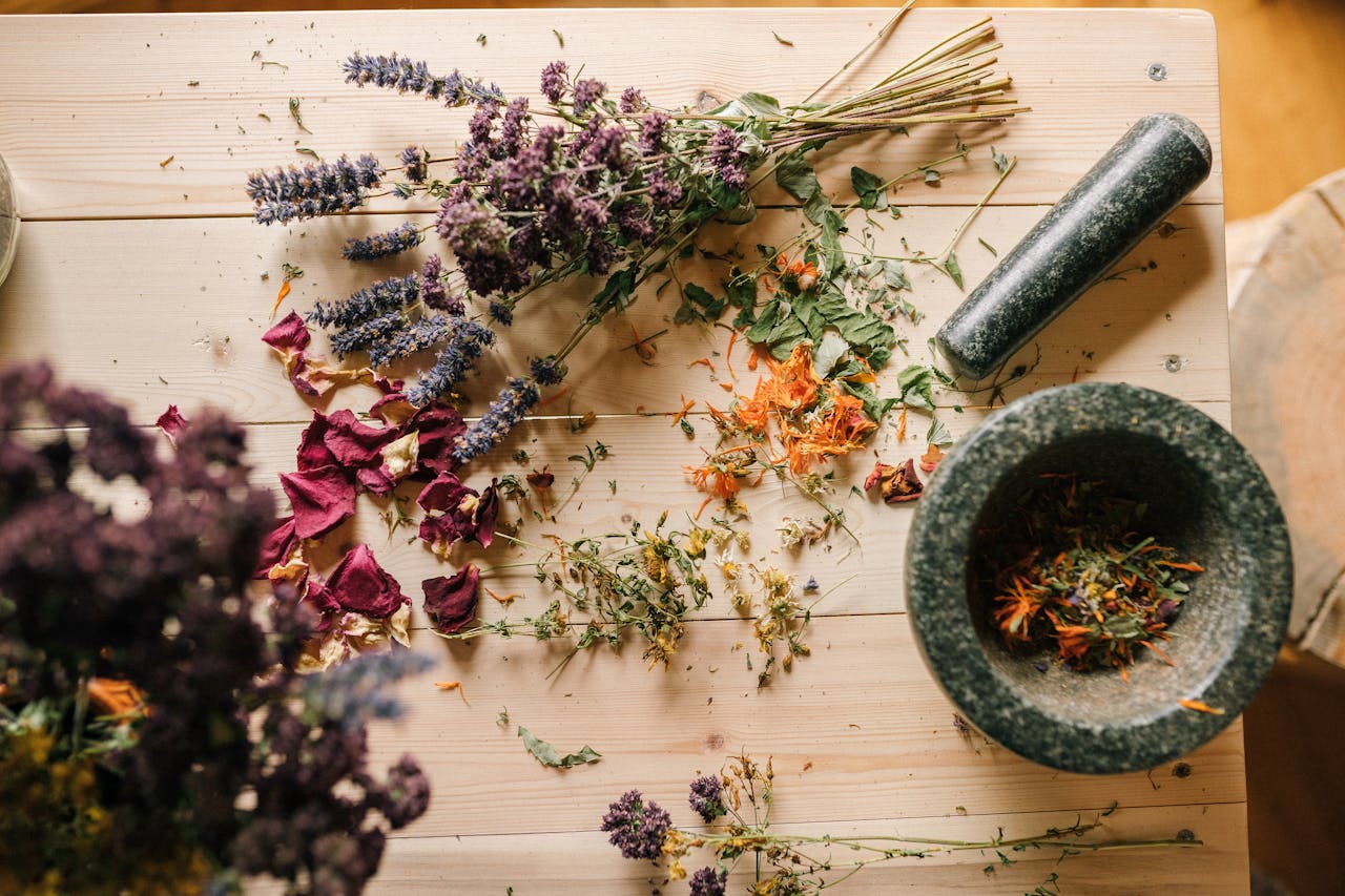 about-01 Flat lay of dried herbs and flowers with a stone mortar and pestle on a rustic wooden table.